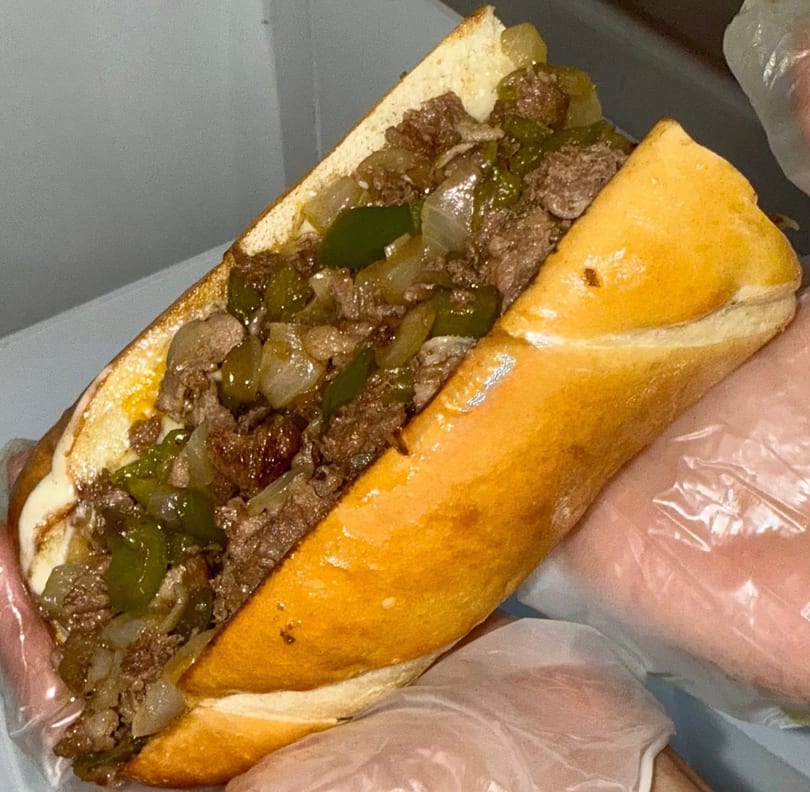 A close-up of a cheesesteak sandwich filled with steak, onions, and green peppers, held by gloved hands.