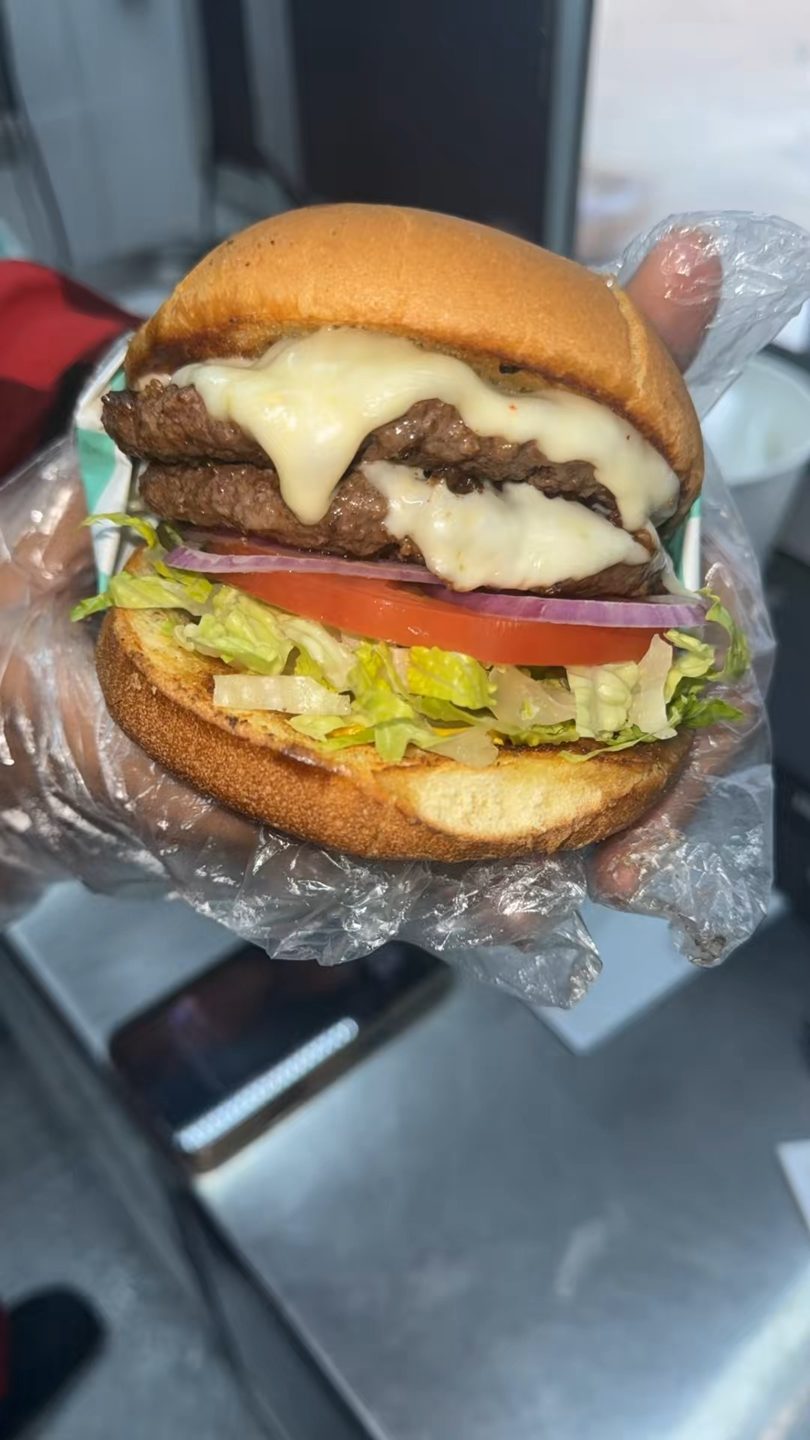 A double cheeseburger with melted cheese, lettuce, tomato, and red onion, held in a plastic-gloved hand.