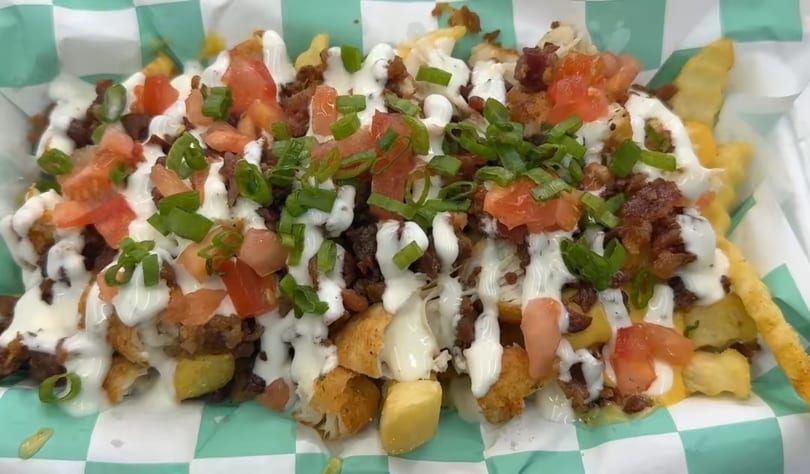 Loaded fries with bacon, tomatoes, green onions, and white sauce in a checkered paper tray.