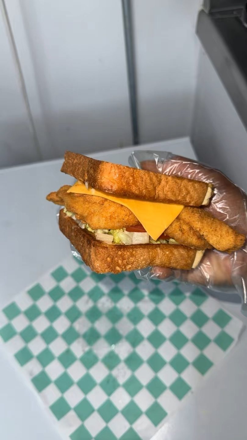 Gloved hands holding a fried fish sandwich with cheese, lettuce, and tomato on toasted bread.