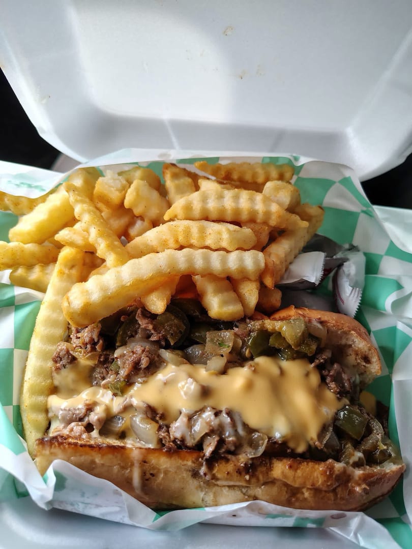 Cheesesteak sandwich with melted cheese, peppers, onions, and a side of crinkle-cut fries.
