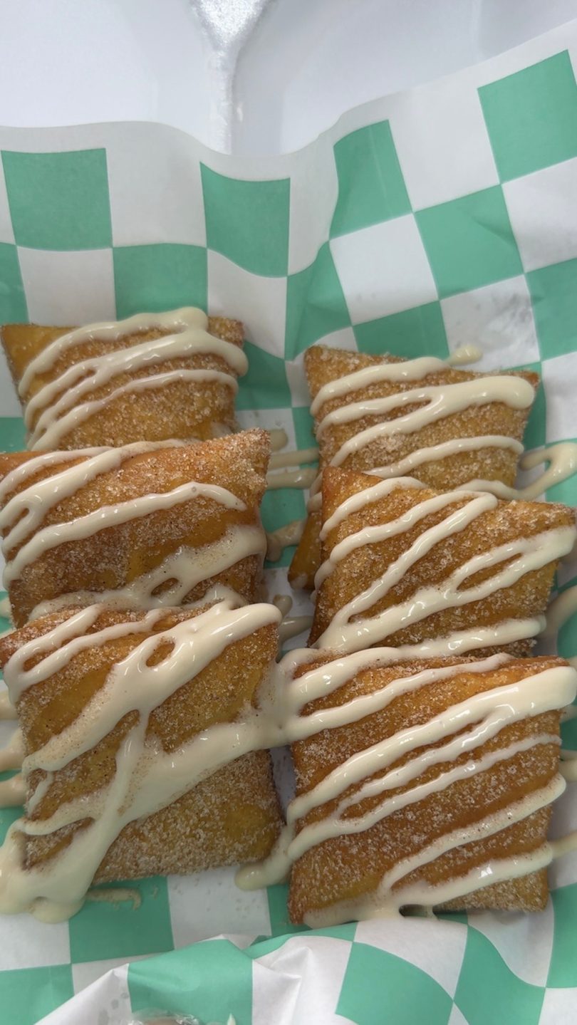 Six golden-brown dessert empanadas, drizzled with white icing, in a green and white checkered paper liner.
